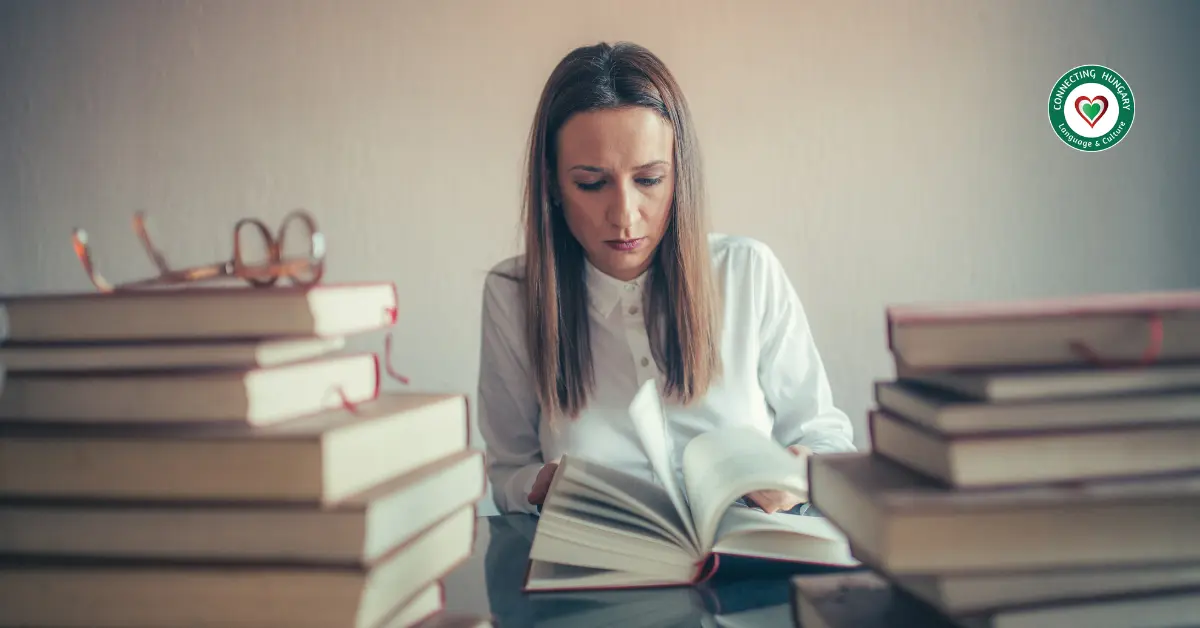 Adult learner reading a book surrounded by stacked books, studying Hungarian at home.